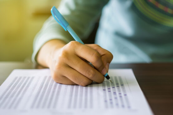 Close up of high school or university student holding a pen writing on answer sheet paper in the examination room. College students answering multiple choice questions test in the testing room in university.